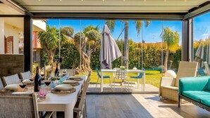 Un'area pranzo all'aperto con un tavolo imbandito per due persone, un divano in vimini e vista su un giardino con palme.
