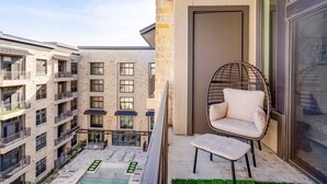 Een balkon met een rieten stoel en poef, uitzicht op een binnenplaats met gebouwen, en een deur aan de rechterkant.