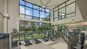 Een fitnessruimte met loopbanden, elliptische trainers en gewichttoestellen, grote ramen en een plafondventilator.