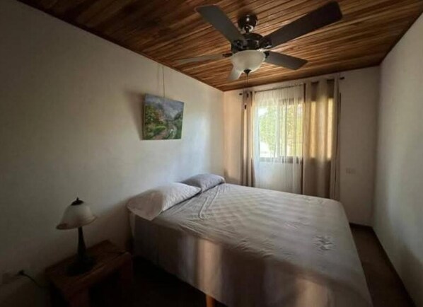 Ein Schlafzimmer mit Bett, Deckenventilator, Nachttisch mit Lampe, einem Wandbild und einem Fenster mit Vorhängen.