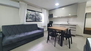 Ein modernes Wohnzimmer mit Sofa, Esstisch, Stühlen, Küchenzeile und einem Fenster mit Blick auf Bäume.