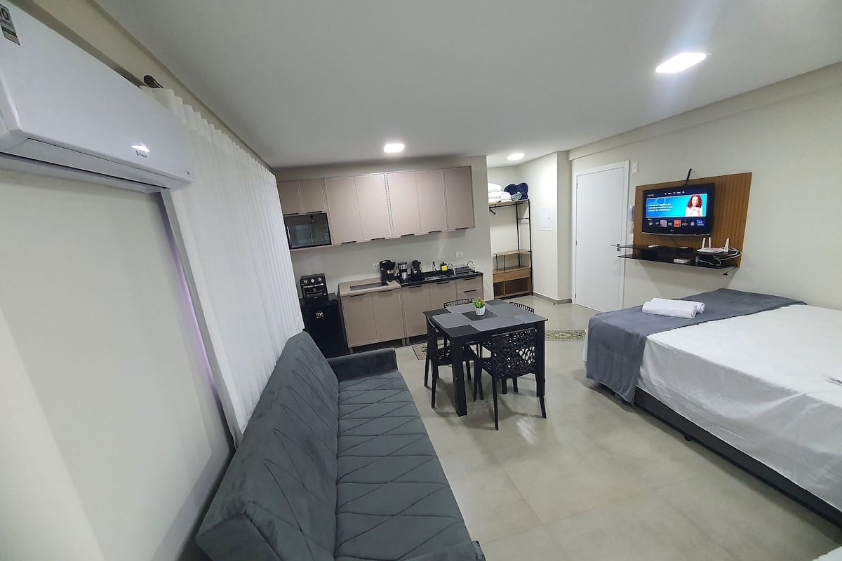 A compact hotel room with a kitchenette, a sofa, a small dining table, a TV, and a bed.