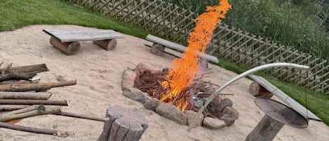 A sandy outdoor area with a fire pit, wooden benches, and a metal grill.