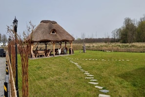 Una construcción con techo de paja, con una mesa y sillas afuera, rodeada por un área cubierta de césped y piedras para pisar.