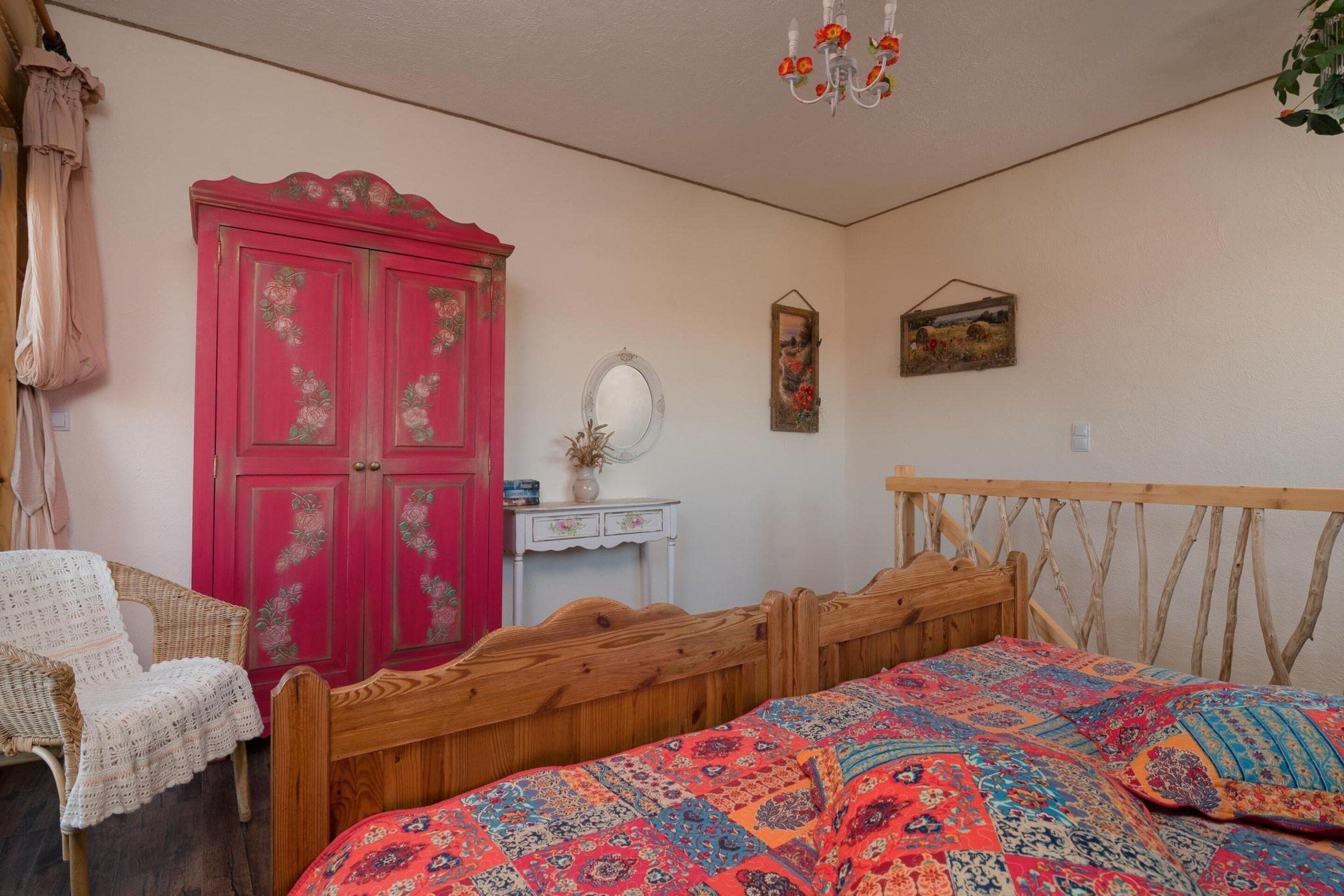 A bedroom with a wooden bed, a red wardrobe, a white bedside table, a hanging mirror, and a wall-mounted picture.