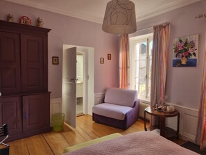 Een kamer met een paarse fauteuil, een bed met een beddengoed met patroon, een houten kast, een groene prullenbak en een raam met gordijnen.