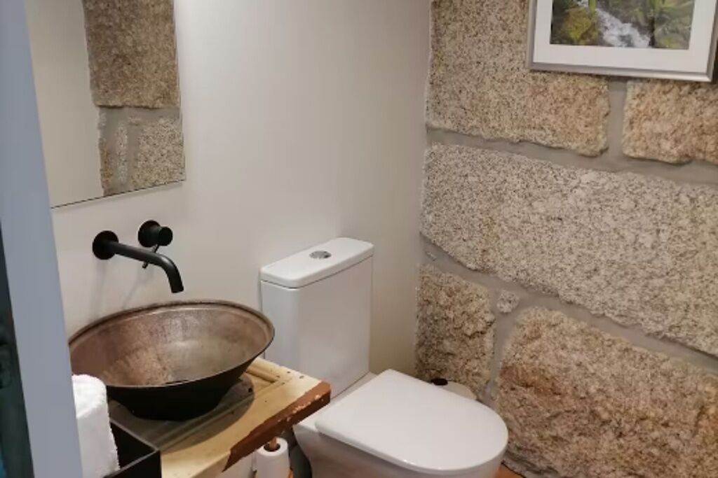 A bathroom with a stone wall, a wooden sink, a white toilet, and a framed picture on the wall.