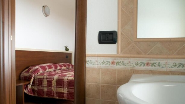 A bedroom with a bed, a bedside table, a wall-mounted light, and a bathroom with a bathtub and a wall-mounted electrical outlet. - Agriturismo Fiorella (Celico)