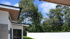 A modern outdoor patio with a round glass table and four chairs, a swimming pool, and a white wall with trees in the background. - Stylish 3BR Villa w/ Pool at Marino Ballena (Uvita)