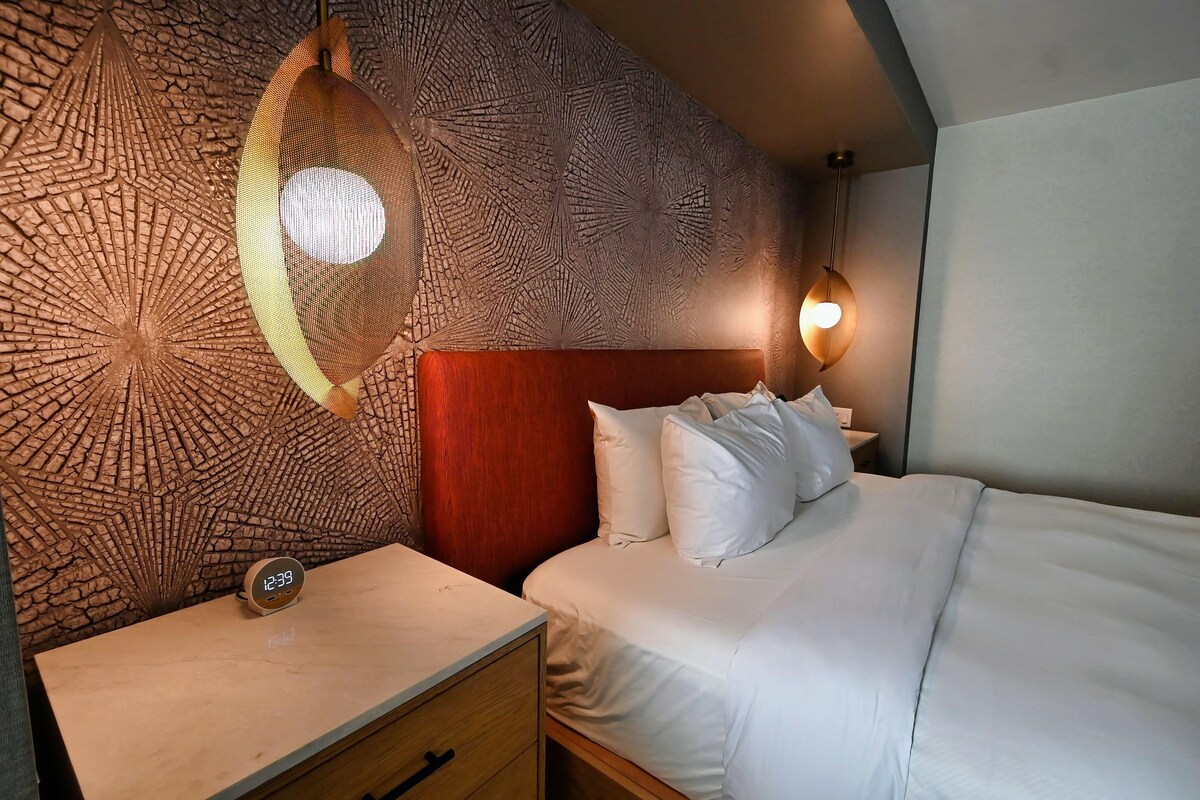 Habitación de hotel con pared texturizada, cabecera de madera, cama con ropa blanca, mesita de noche con un reloj y una lámpara colgante.