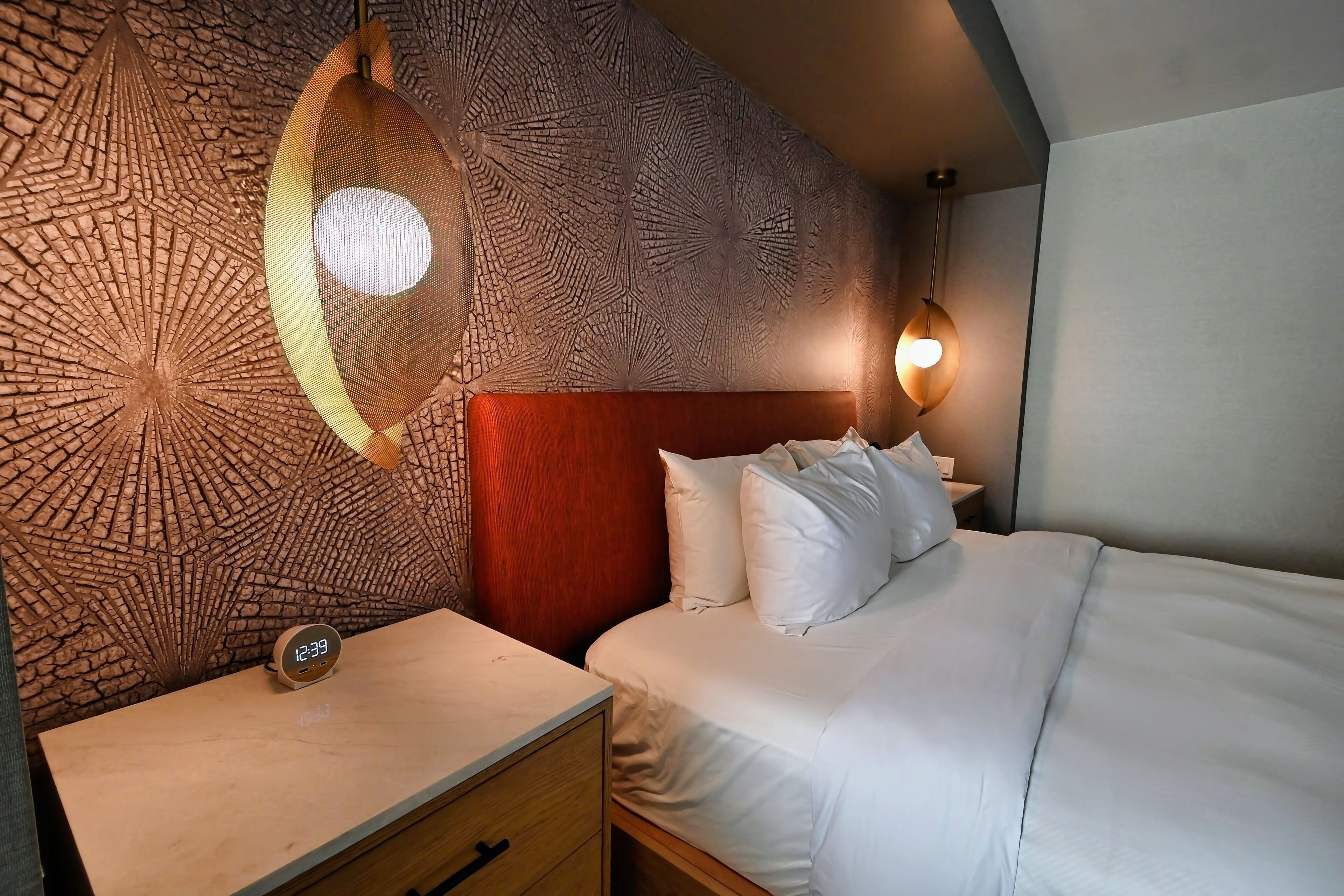A hotel room with a textured wall, a wooden headboard, a bed with white bedding, a bedside table with a clock, and a hanging light fixture.