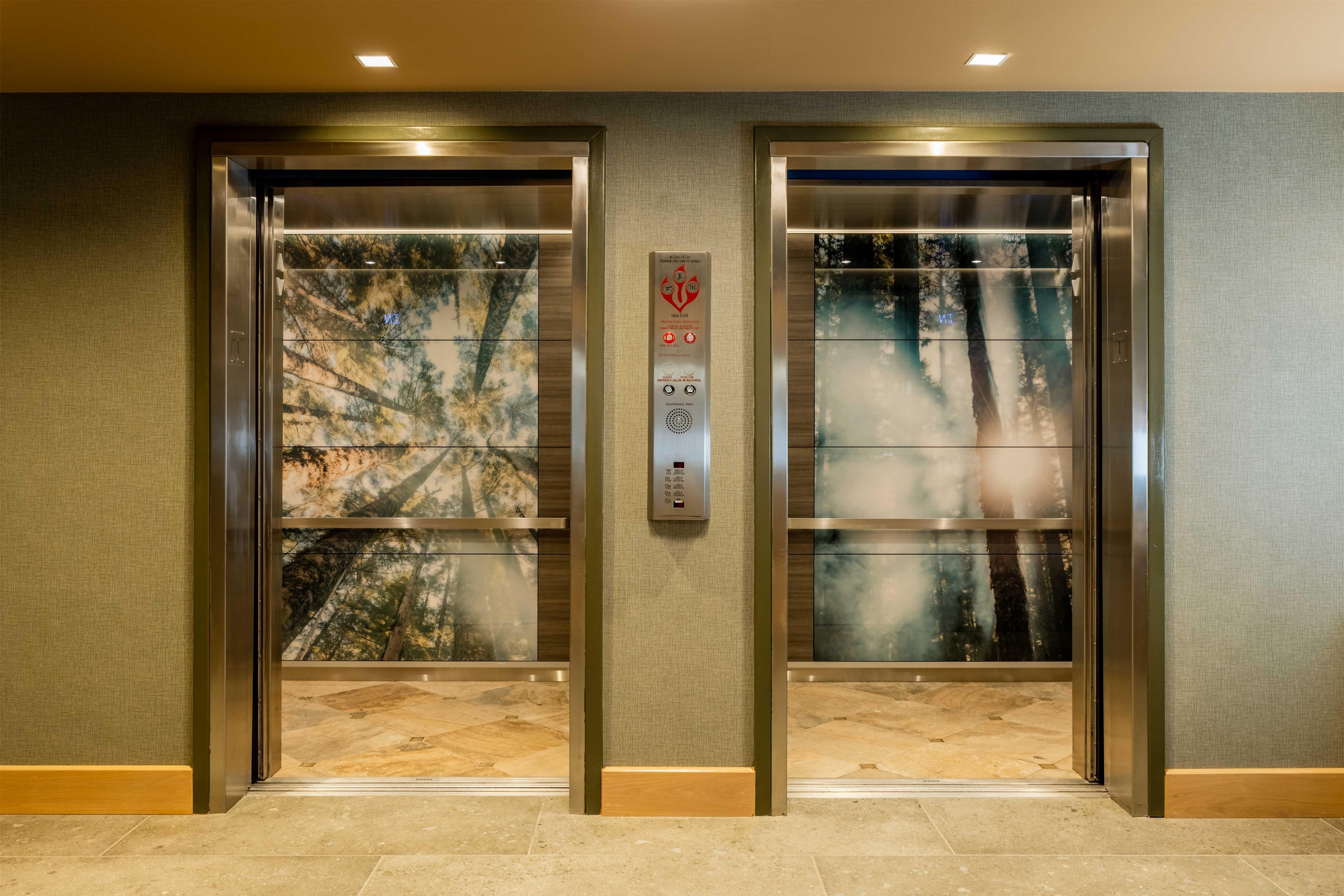 Two elevator doors with a forest view, a control panel with emergency instructions, and a textured wall.