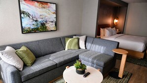 A modern hotel room with a grey sofa, a round coffee table with a plant, a bed with white bedding, and a painting on the wall. - The Mindaro, part of JdV by Hyatt (Portland)