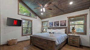 Ein Schlafzimmer mit Deckenventilator aus Holz, einem Bett mit grauer Bettdecke, einem an der Wand befestigten Fernseher und einem Fenster mit Jalousien.