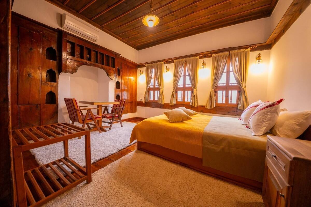 Une pièce confortable avec un lit, une table en bois entourée de chaises et une fenêtre avec des rideaux.