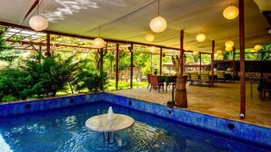 Une piscine extérieure agrémentée d’une fontaine, entourée de tables et de chaises en bois, et située sous une zone abritée illuminée par des lanternes suspendues.