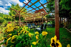 Un área de estar al aire libre techada con sillas y mesas, rodeada de follaje y flores.