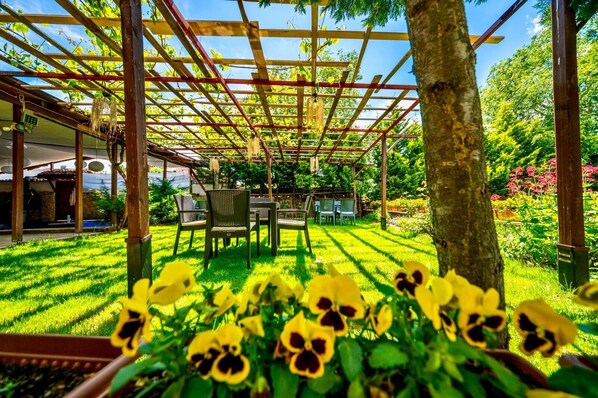 Un espace extérieur aménagé, abrité et doté de chaises et de tables, entouré d’une végétation luxuriante et de plantes en fleurs.