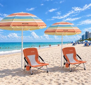 Une plage avec deux chaises de plage et des parasols, un ciel dégagé et, en arrière-plan, la silhouette d’une ville.