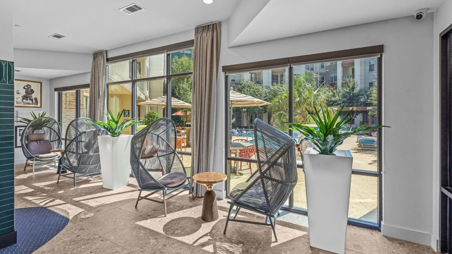 Quarto de hotel moderno com grandes janelas, cadeiras de vime, uma pequena mesa redonda e plantas em vasos.