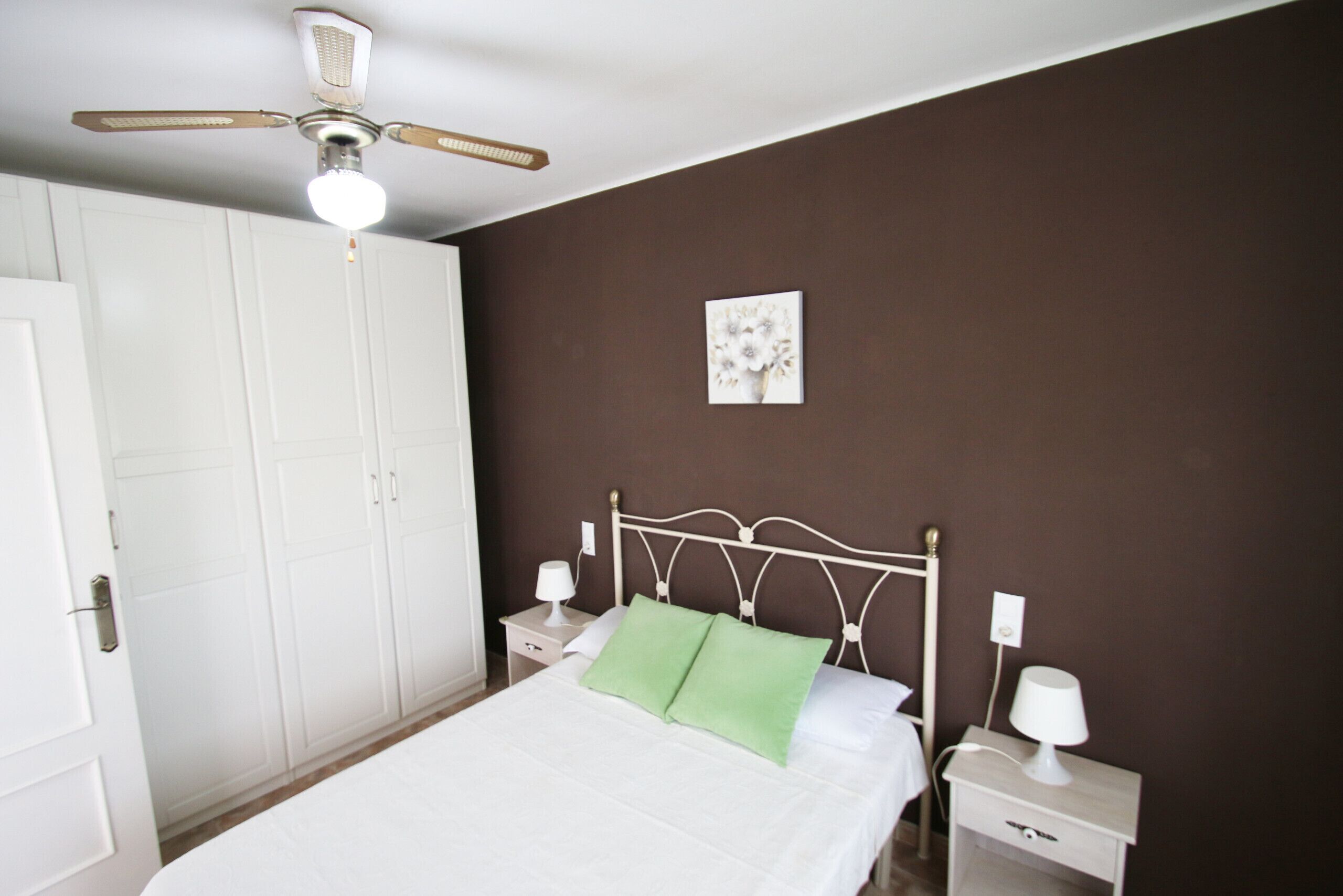 Une chambre à coucher comprenant un lit, des tables de chevet, un ventilateur de plafond, une armoire et un tableau accroché au mur.