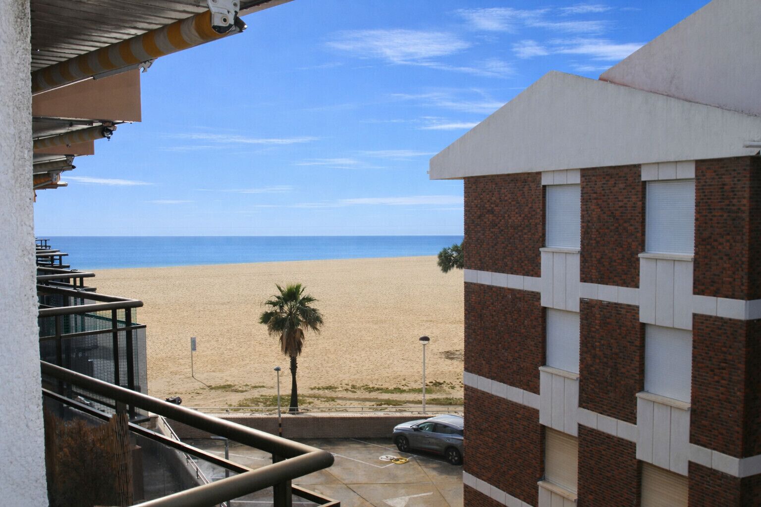 A beach view from a balcony overlooking a sandy beach, a palm tree, and a building with a brick facade.