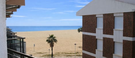 A beach view from a balcony overlooking a sandy beach, a palm tree, and a building with a brick facade.