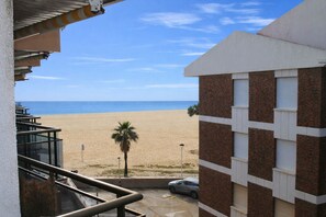 A beach view from a balcony overlooking a sandy beach, a palm tree, and a building with a brick facade. - Apartment Membling Cambrils (Cambrils)