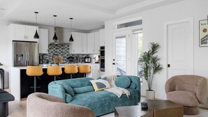 Une cuisine moderne avec des meubles blancs, un réfrigérateur en acier inoxydable et un comptoir noir. On y trouve trois tabourets de bar jaunes et un canapé turquoise agrémenté d’un plaid.