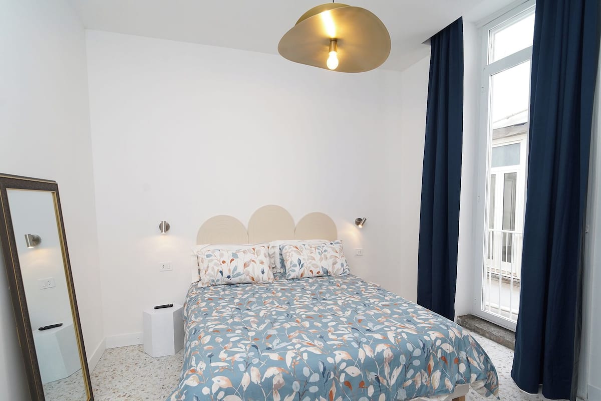 A bedroom with a bed, a mirror, a bedside table, and a window with curtains.