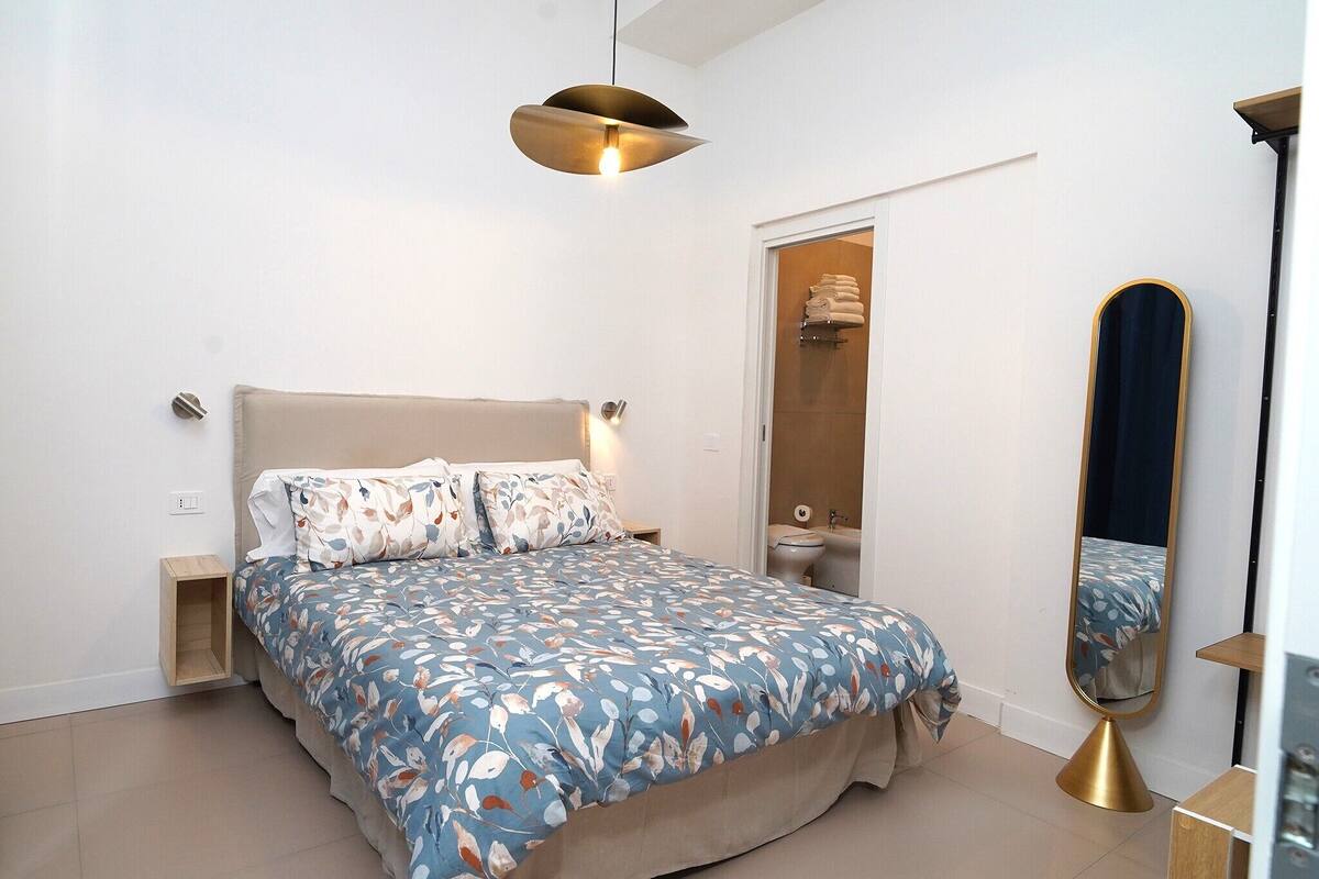 A bedroom with a bed, bedside tables, a hanging light fixture, a mirror, and a door leading to a bathroom.