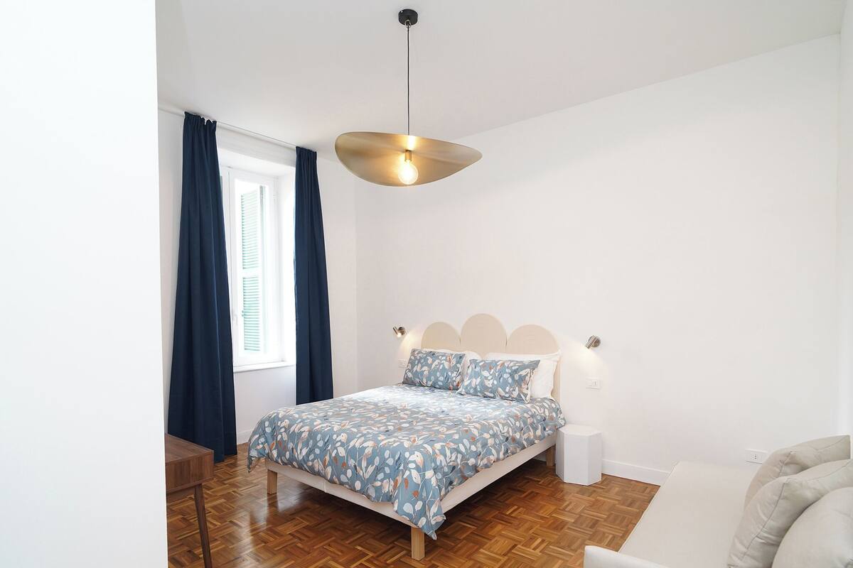 A bedroom with a bed, a bedside table, a chair, a wooden floor, a window with curtains, and a pendant light.