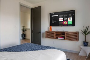 A bedroom with a bed, a TV mounted on the wall displaying streaming services, a wooden shelf with books, and a potted plant. - Ultra-Spacious Boutique Retreat in Downtown Dallas (Dallas)