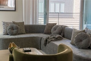Ein modernes Wohnzimmer mit einem grauen Ecksofa, mehreren Kissen, einem kleinen Tisch mit einer Schachtel und einem Fenster mit Blick auf Gebäude.