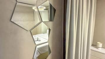 Une pièce avec un miroir au motif géométrique sur le mur, un lit recouvert de draps blancs, une table de chevet avec une lampe et une fenêtre avec des rideaux.