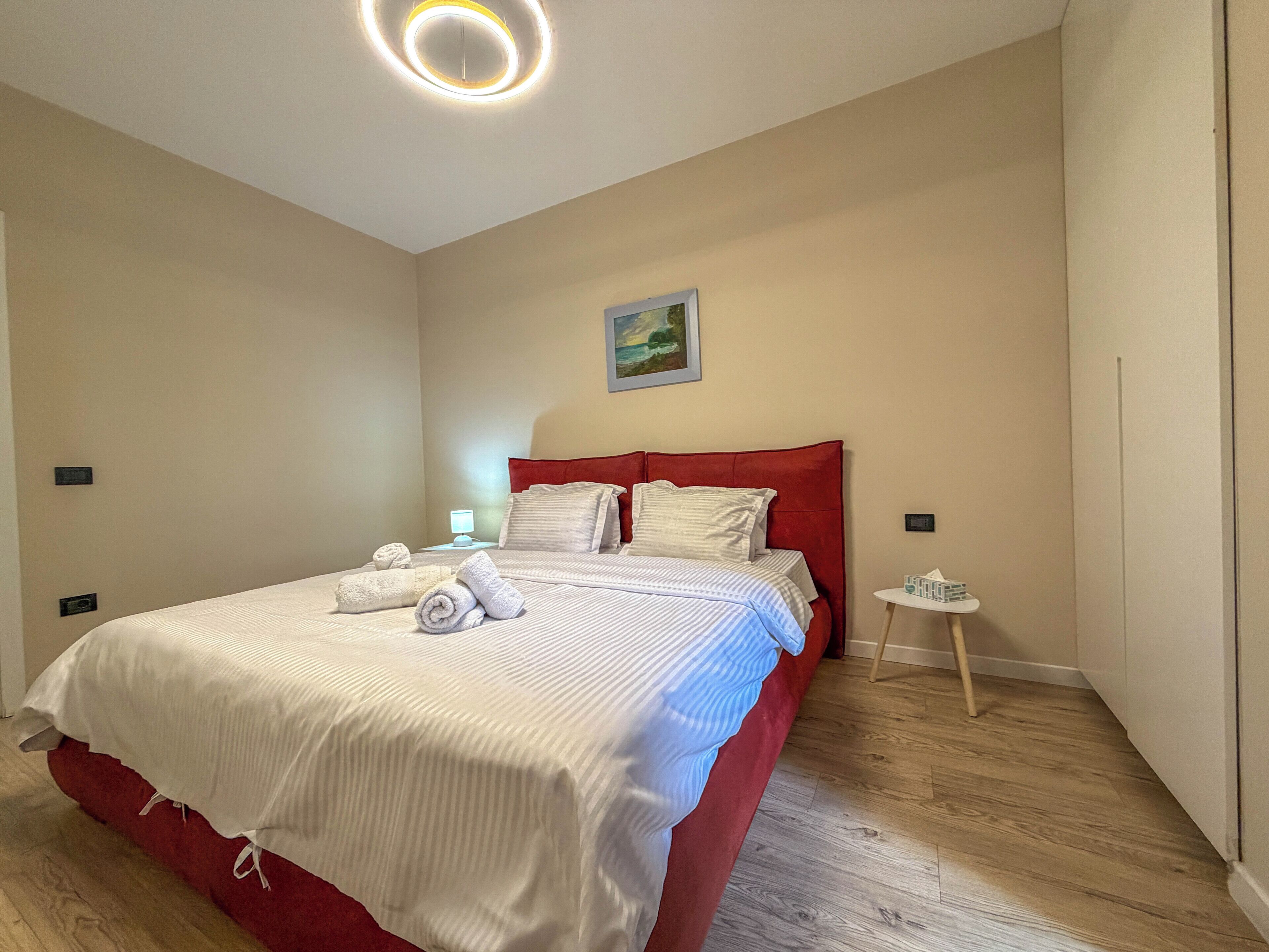 A neatly made bed with white linens and rolled towels, a red headboard, a bedside table with a lamp, and a small round table with a tissue box.