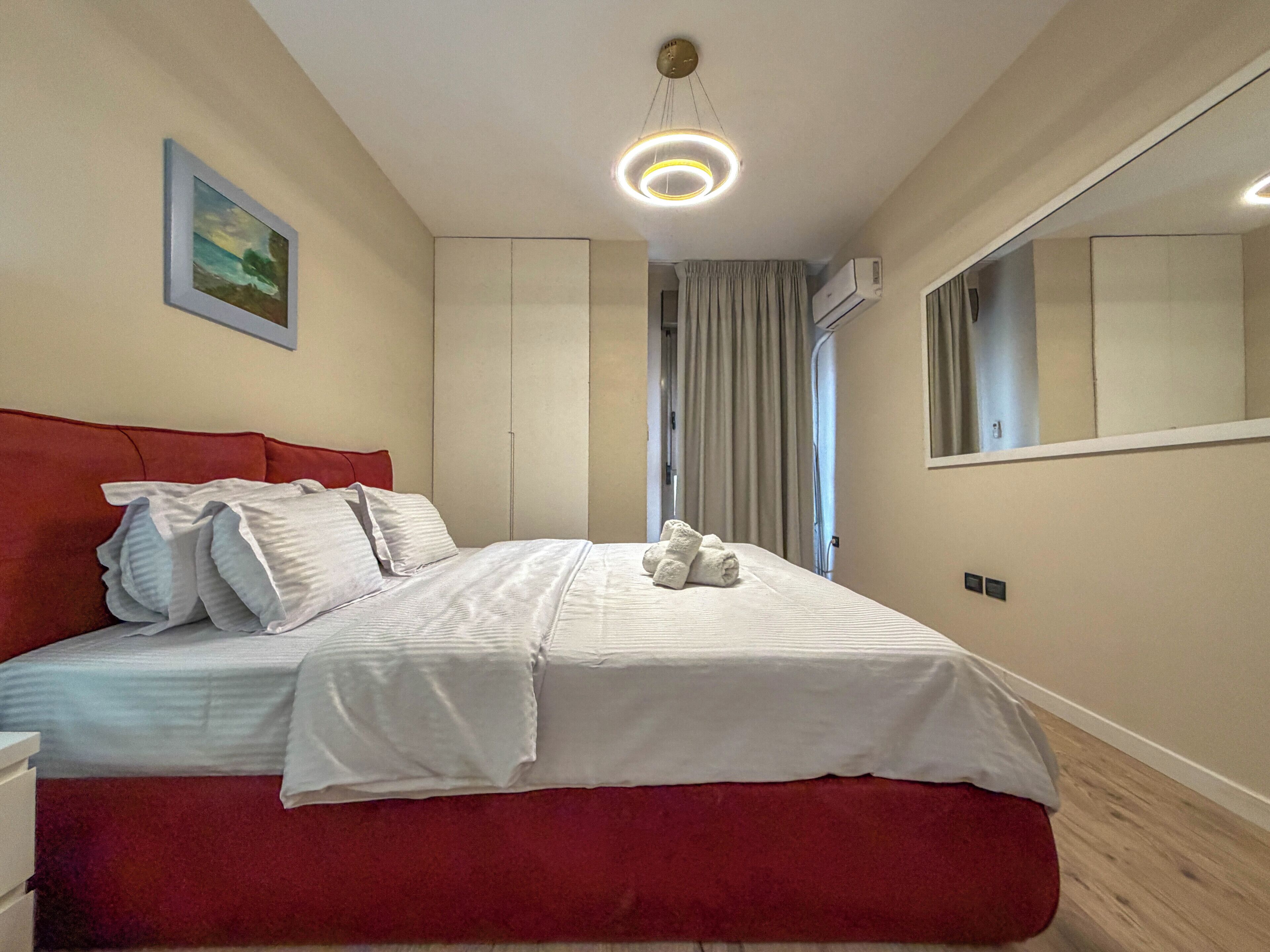 A bedroom with a bed, a red headboard, a painting, a ceiling light, and an air conditioning unit.