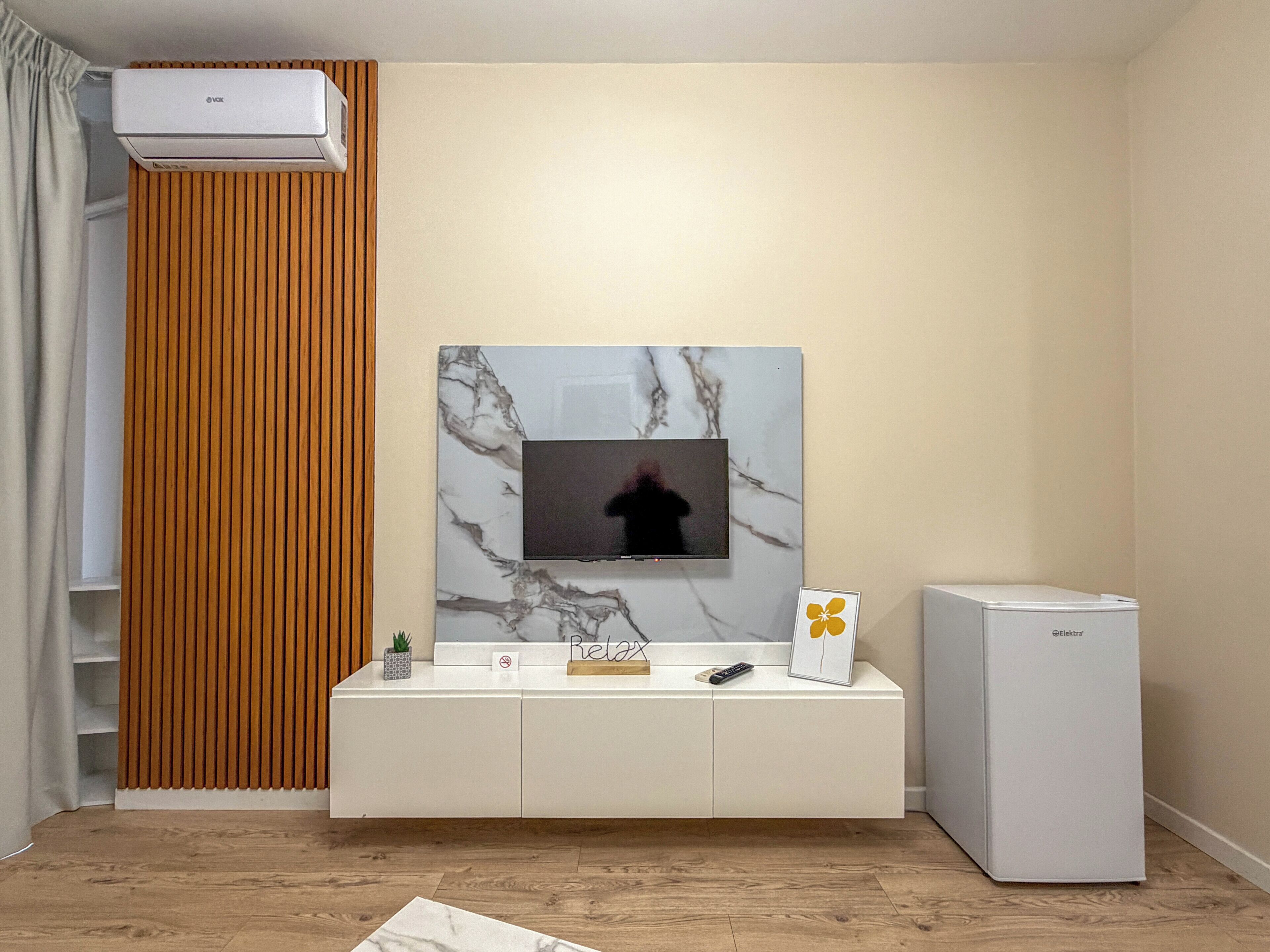 Una sala de estar moderna con un televisor montado, un mueble blanco, un refrigerador pequeño y una pared de paneles de madera.