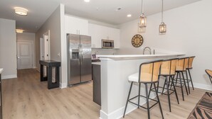 A modern kitchen with a central island, bar stools, stainless steel appliances, and pendant lighting. - New Coastal Condo - Walk to Shops + Bike to Beach (Rehoboth Beach)