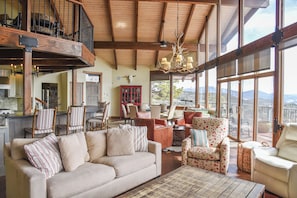 A spacious living area with a high ceiling, wooden beams, a large window with a mountain view, a fireplace, and a cozy seating arrangement.