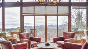 Ein Zimmer mit Ledersesseln um einen runden Tisch, großen Fenstern mit Blick auf die Berge und einer Holzbalkendecke.