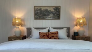 A neatly made bed with white linens, two bedside tables with lamps, and a framed landscape painting on the wall.