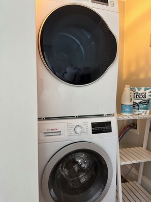 Un lave-linge Bosch à chargement frontal, un sèche-linge placé au-dessus et une étagère contenant des produits de lessive.