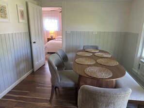 Un coin repas avec une table en bois, six sets de table en fibres tressées, des chaises à l’assise rembourrée de couleur grise et une vue sur une chambre à coucher avec un lit et des rideaux.
