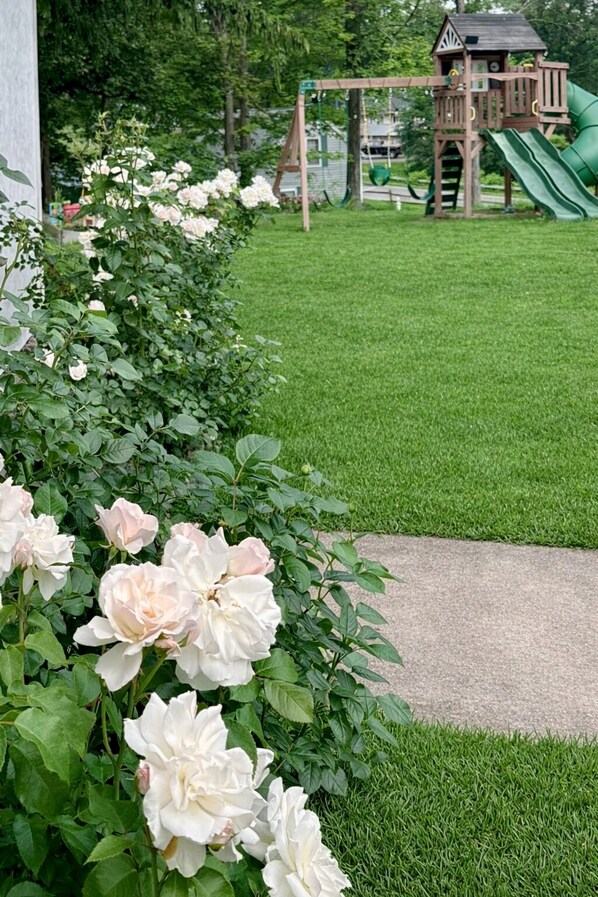 Un jardin avec des rosiers, une pelouse verdoyante et une structure de jeu en bois avec un toboggan.