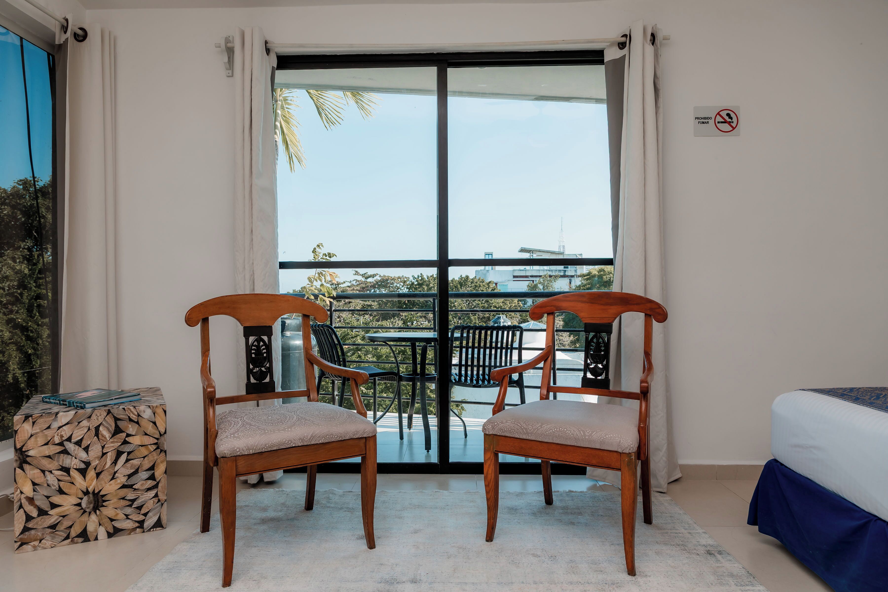 Habitación con balcón, dos sillas de madera, una cama, una mesita auxiliar y vistas al exterior.