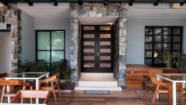 Une entrée moderne avec un mur en pierre, une porte noire et un paillasson. À l’extérieur, on trouve des tables et des chaises en bois.