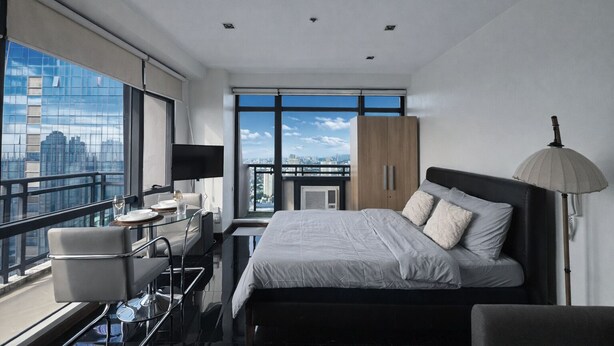 A modern hotel room with a large bed, a desk with a chair, a television, and a view of the cityscape through large windows. - Executive Studio w/ Sunset View - Makati CBD (Makati)