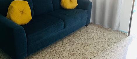 A room with a dark blue sofa, two yellow cushions, a ceiling light fixture, and a window with a curtain.