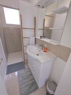 Ein Badezimmer mit einem weißen Waschbecken, einem Spiegel und einer Dusche. Auf dem Waschbecken liegen Pflegeprodukte und es gibt einen Handtuchhalter.